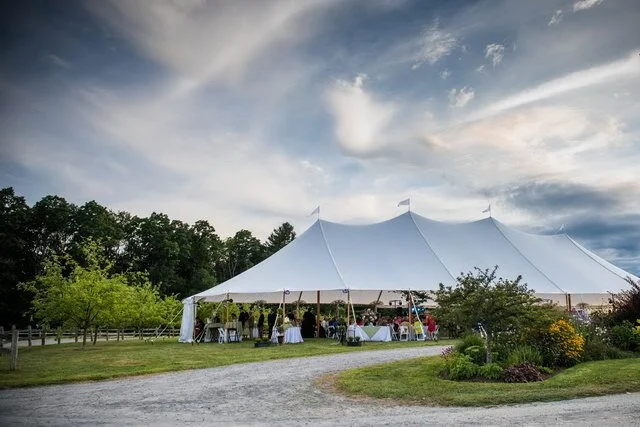Large white event tent set up outdoors with people gathered underneath, surrounded by green trees and a garden, under a partly cloudy sky.