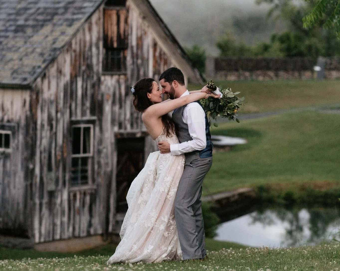 A newlywed couple embracing outdoors near a rustic wooden barn, with greenery and a pond in the background.
