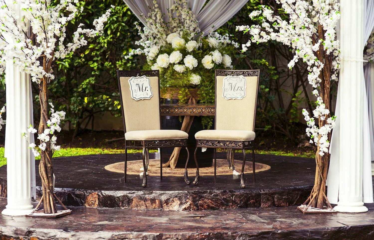 Wedding ceremony setup with two beige chairs labeled Mr. and Mrs., surrounded by white floral arrangements and greenery on a small wooden stage.